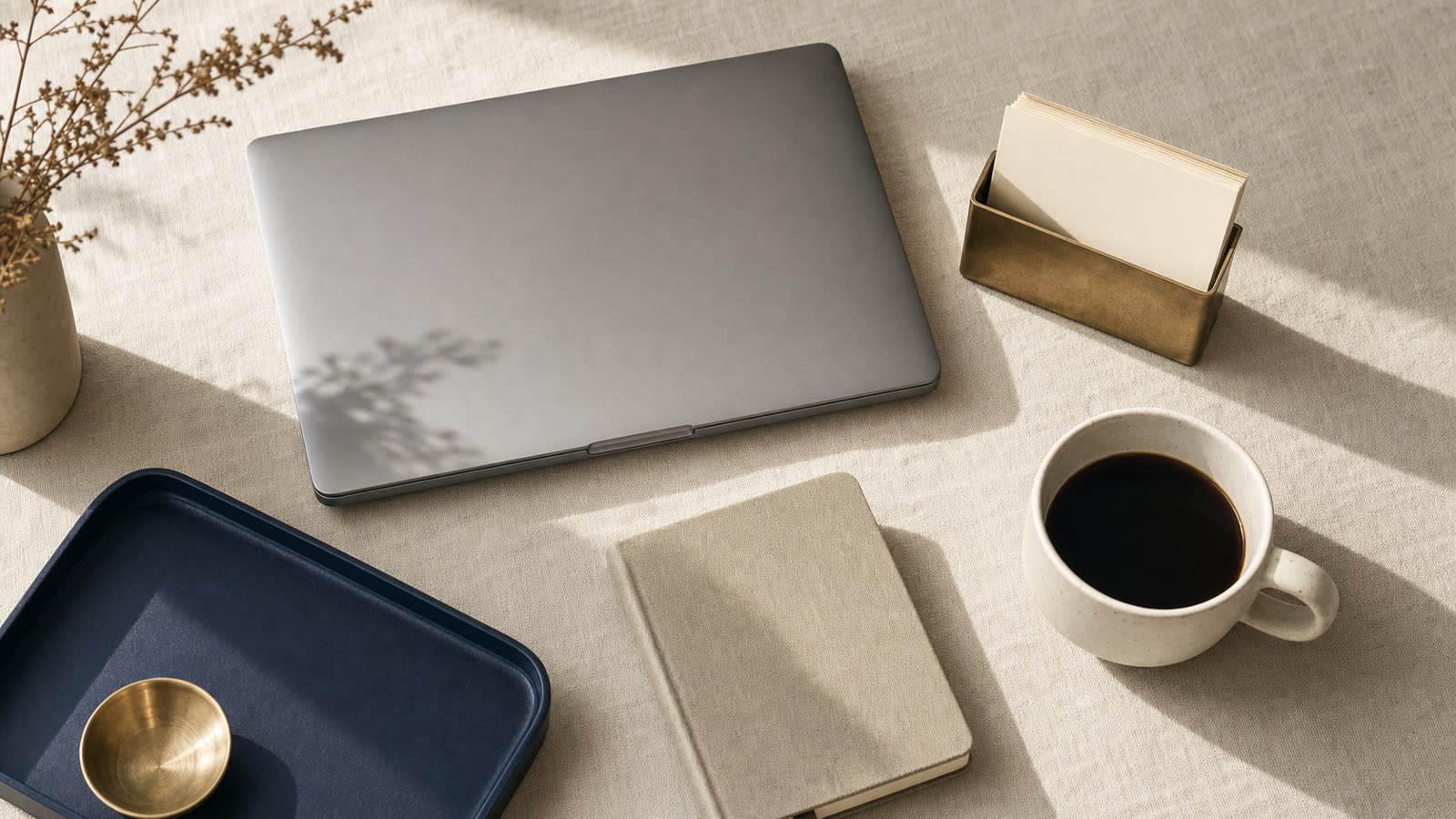 Overhead view of a slim laptop, blank cream business cards in a brass holder, and coffee on warm linen